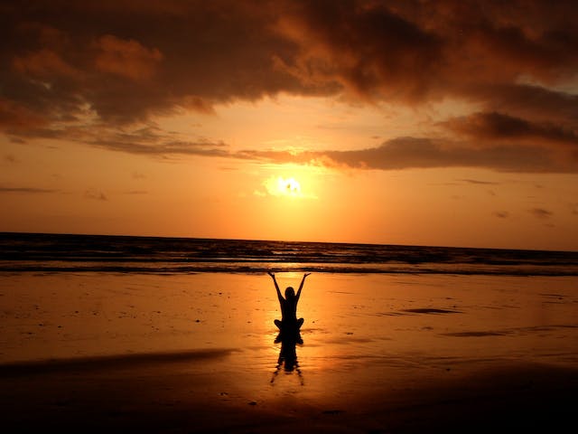 mujer con brazos extendidos hacia el sol sentada sobre la arena en playa en el atardecer